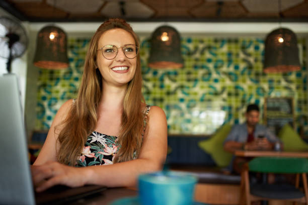 Beautiful young positive female entrepreneur looking away with smile, while typing on laptop during work session in trendy coffee shop restaurant serving fair-trade organic coffee