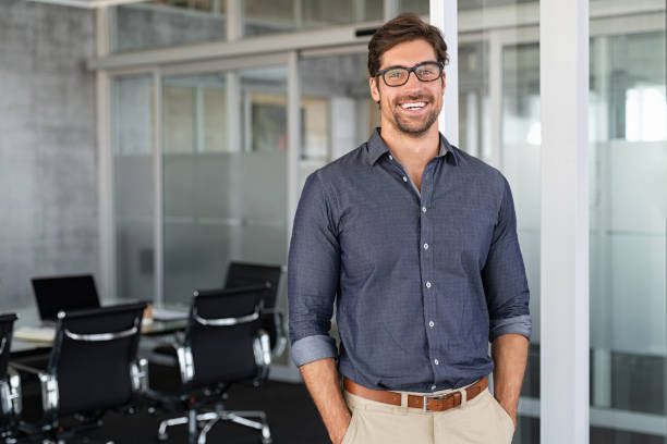 Portrait of young businessman wearing eyeglasses and standing outside conference room. Portrait of happy business man wearing spectacles and looking at camera with copy space. Satisfied proud man feeling confident in a modern office.